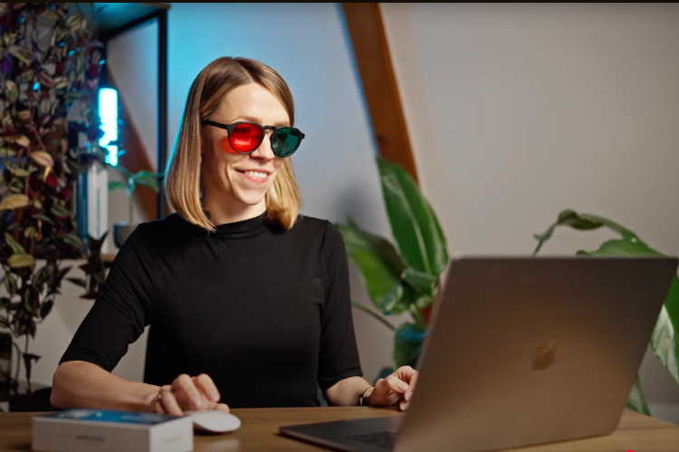 A woman with red-green glasses sitting behind the desk looking into computer.