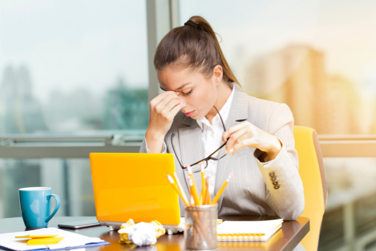 Woman in the office, sitting behind the desk, holding the top of her nose with right hand.