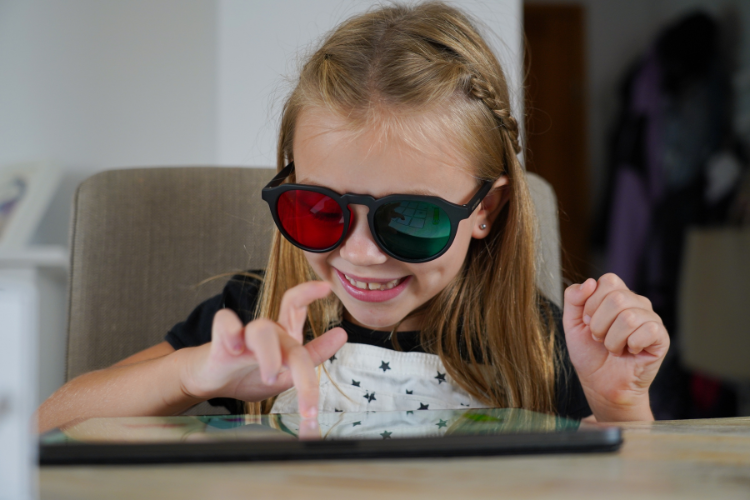 Happy young girl with red-green glasses sitting on the chair at the table, playing games on a tablet with her right hand