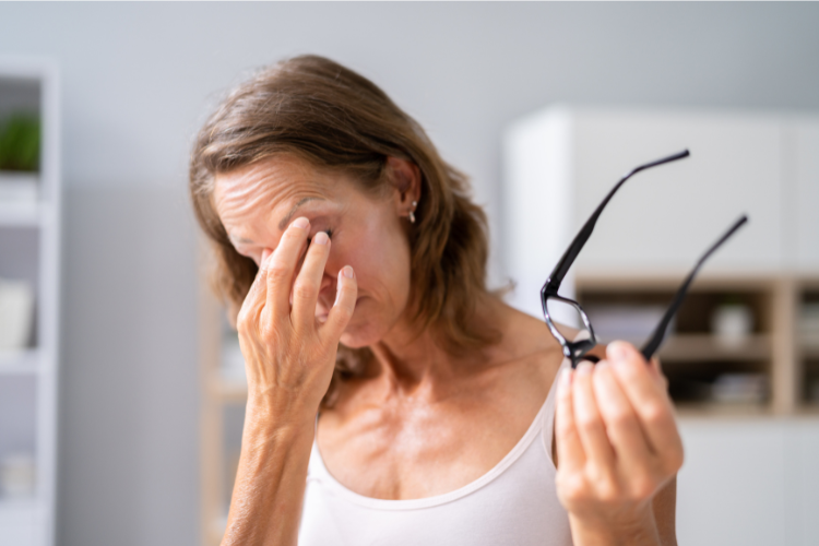 Woman rubbing irritated eyes while holding glasses, representing eye strain and poor sleep